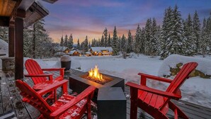Una terraza con sillas Adirondack rojas y un brasero, rodeada de árboles y casas cubiertas de nieve.