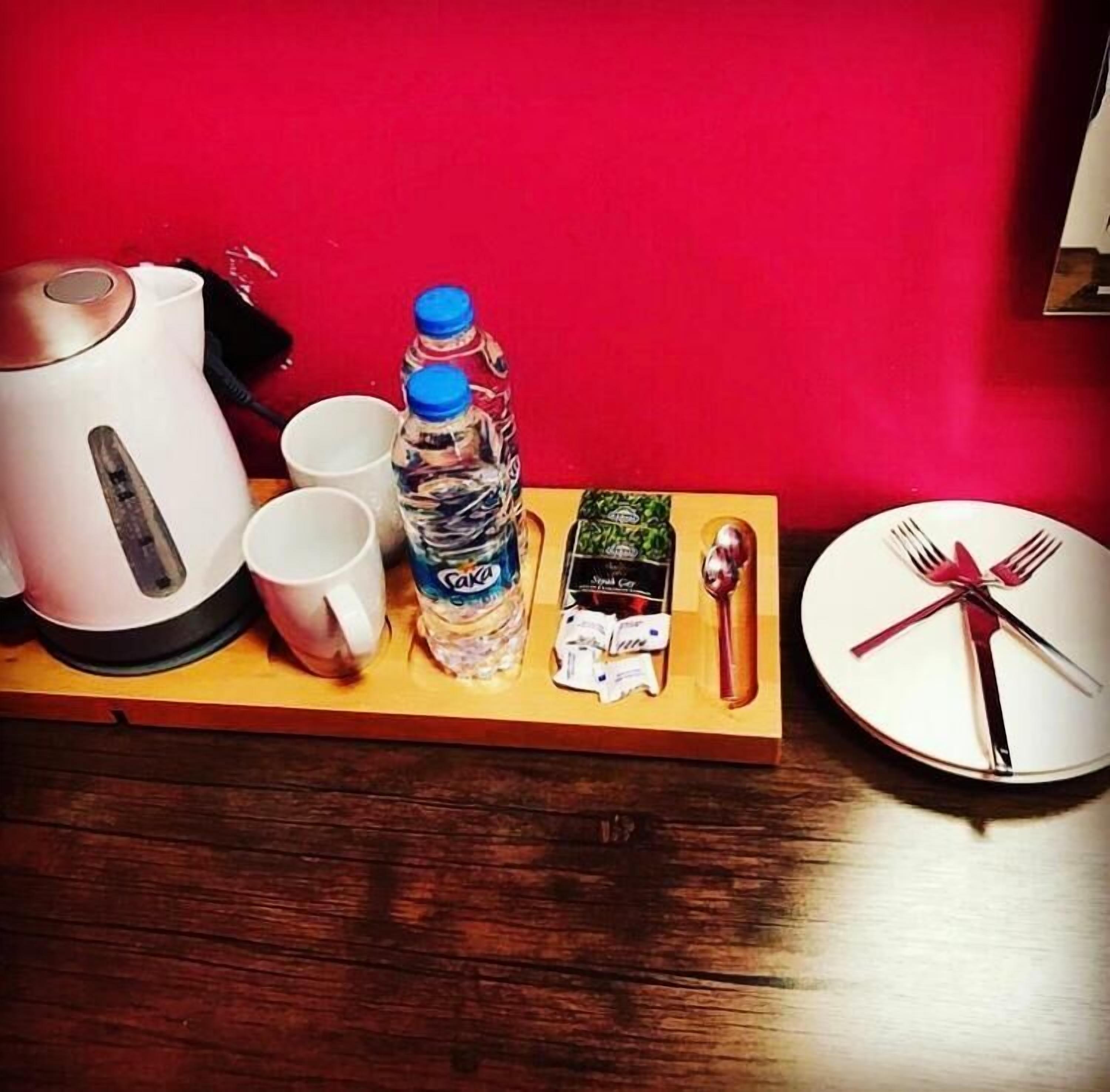 A wooden tray with a teapot, two cups, a water bottle, a box of snacks, and a plate with cutlery.