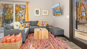 Ein modernes Wohnzimmer mit einem grauen Ecksofa, einem Holztisch und einem farbenfrohen Teppich. An der Wand befinden sich gerahmte Bilder und ein großes Fenster mit Vorhängen.