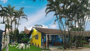 A yellow building with a sloped roof, surrounded by palm trees and a small garden with white chairs. - Pousada Chalé do Beija-flor  (Ubatuba)
