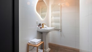 A modern bathroom with a pedestal sink, a wooden stool with towels, a mirror with a circular frame, and a window with blinds. - This hotel is a 1 bedroom(s), 1 bathrooms, located in Delft, Zuid-Holland. (Delft)