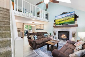 A spacious living room with a fireplace, a large flat-screen TV mounted above it, and a balcony with a wooden railing. - Cozy Bear Lair - Arrowhead Lake (Pocono Lake)