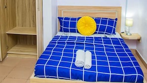 Ein Bett mit einer blau-weißen karierten Tagesdecke, einem gelben Kopfkissen und zwei aufgerollten Handtüchern. Es gibt einen Holzschrank mit einem Spiegel und ein gerahmtes Bild an der Wand.