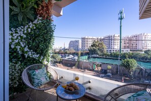 Un balcon avec des fauteuils en osier, une table avec des croissants et du jus d’orange, et une vue sur des immeubles et une rue.