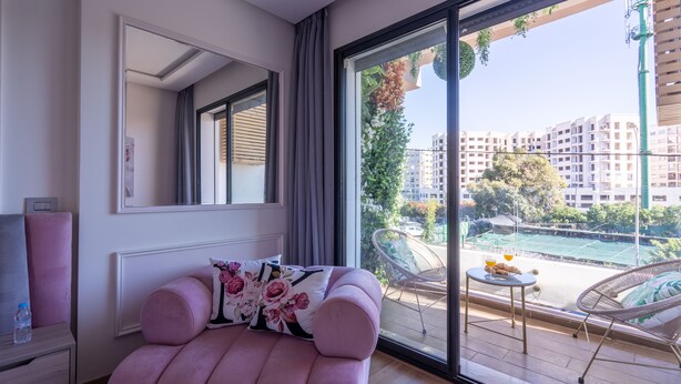 Ein modernes Schlafzimmer mit einem großen Fenster, einem rosa Sofa, einem kleinen Tisch mit Orangensaft und Blick auf Stadt und Grünflächen.