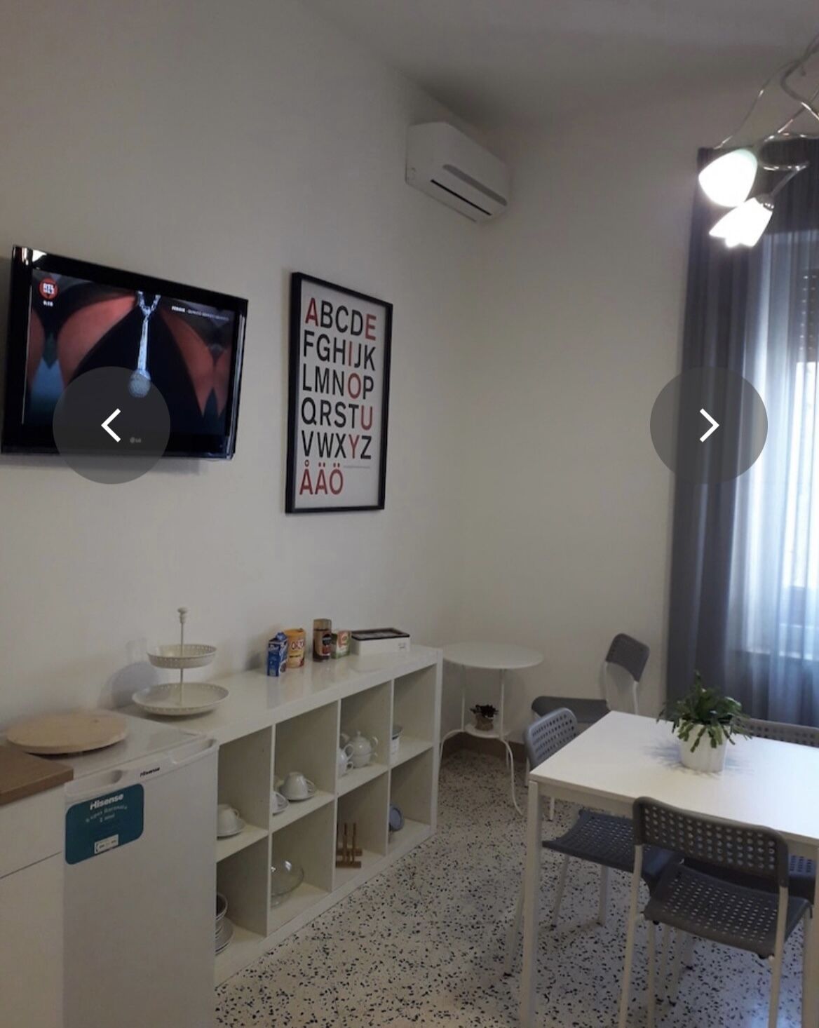 A modern, well-lit room with a flat-screen TV, a wall-mounted air conditioner, a white shelving unit with kitchenware, a small table with chairs, and a potted plant.
