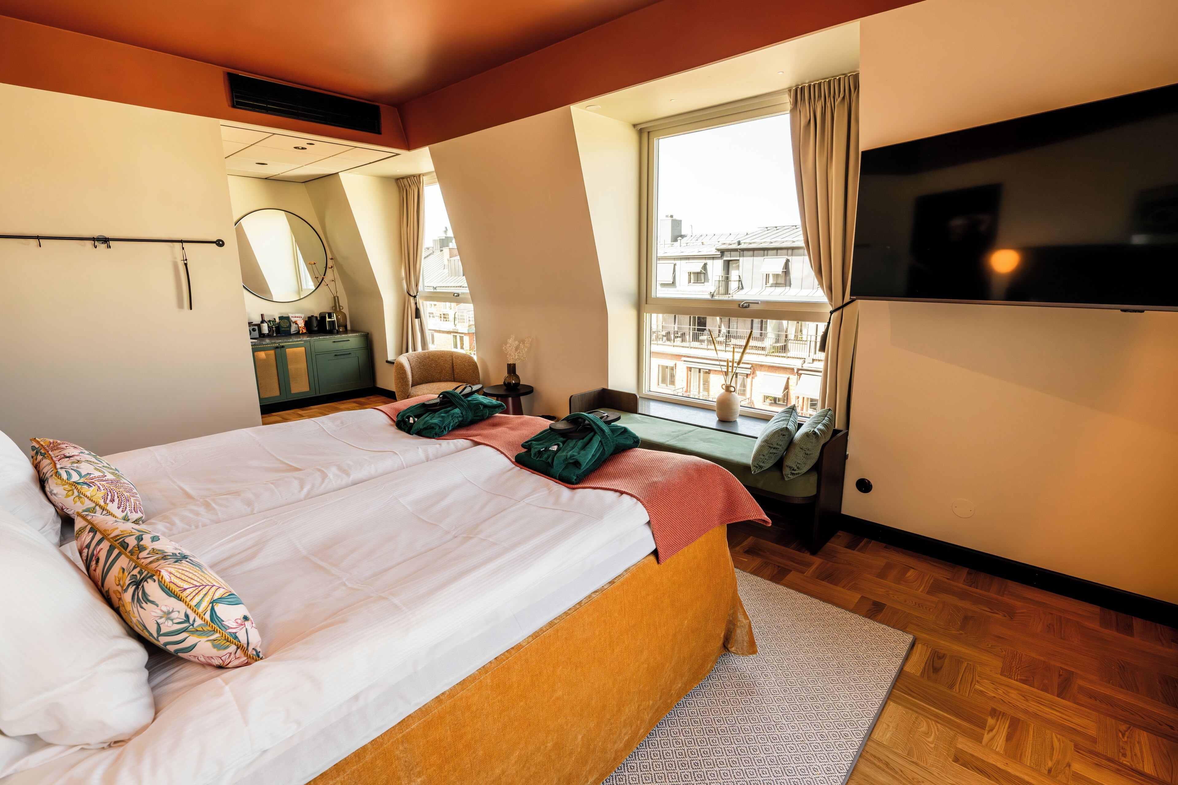 Une chambre avec un grand lit, une télévision, une fenêtre avec des rideaux, un bureau avec une chaise et une vue sur les bâtiments à l’extérieur.