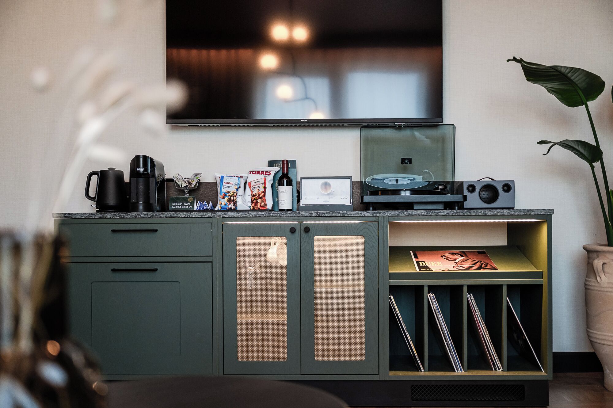 Une cuisine moderne avec un téléviseur à écran plat fixé au-dessus d’un plan de travail, une machine à café, une bouteille de vin et un tourne-disque.