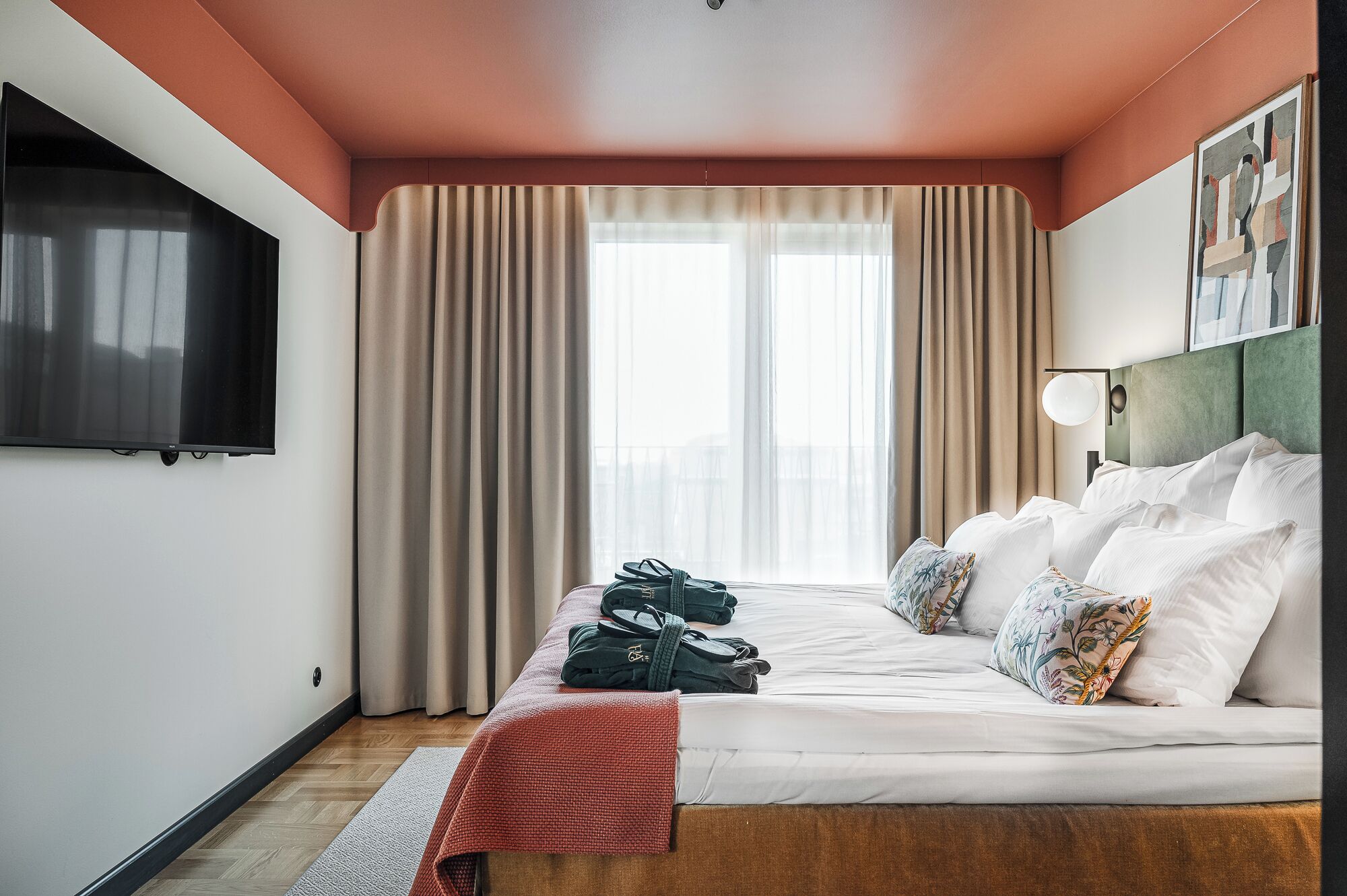 A hotel room with a bed, a flat-screen TV, a headboard, and a window with curtains.