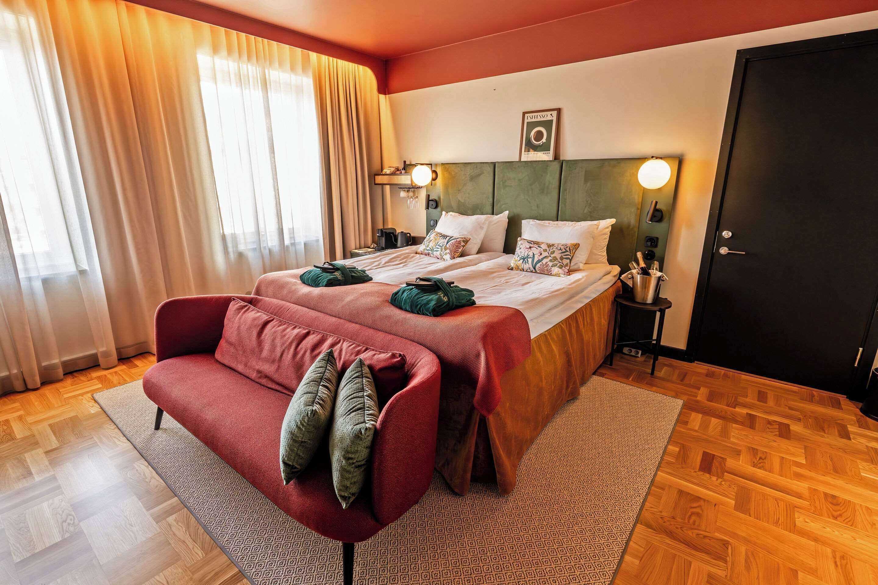 A hotel room with a bed, a red sofa, a wooden floor, and a large window with curtains.