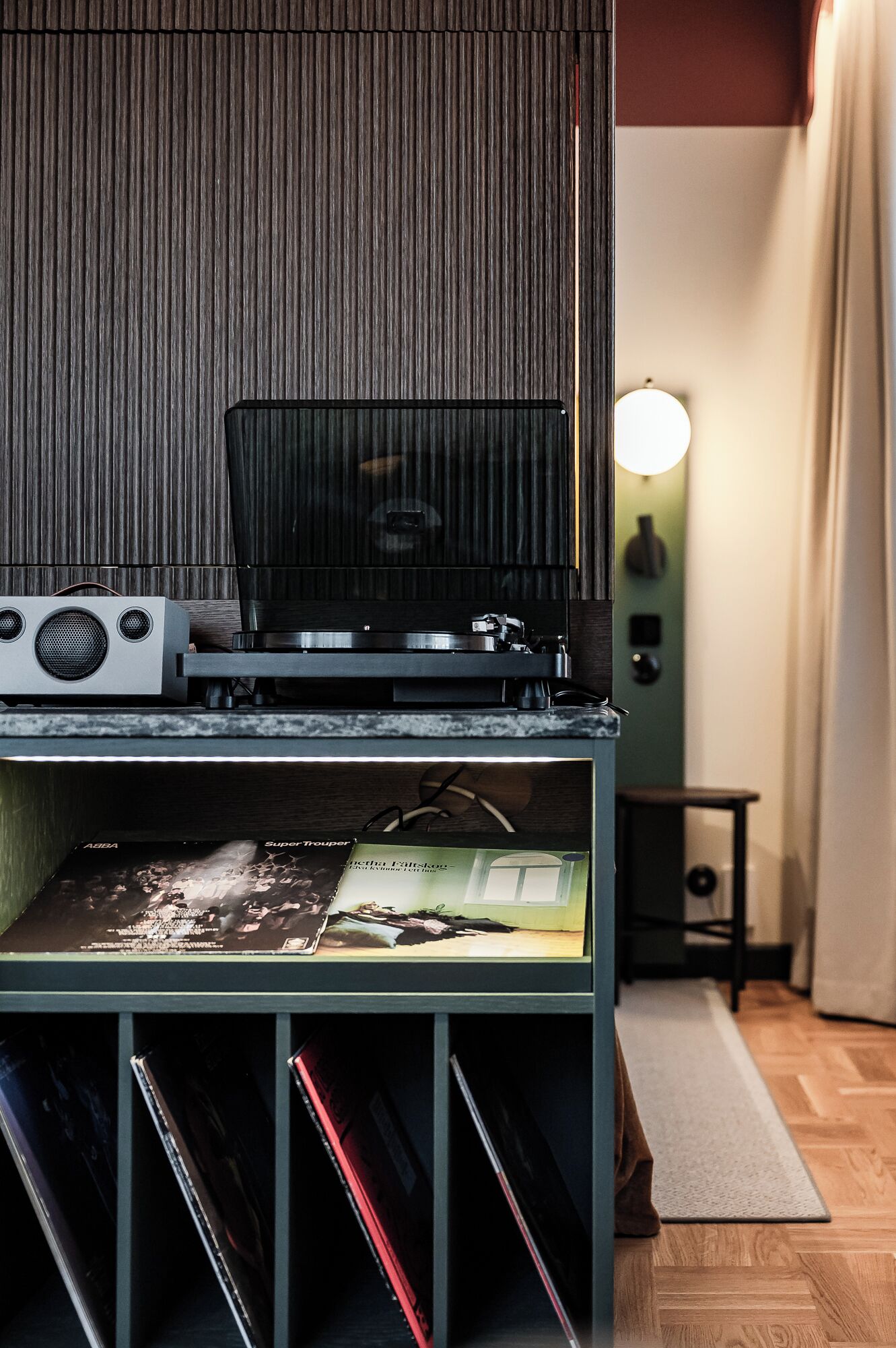 A room with a vinyl record player on a shelf, a small table with a lamp, and a window with curtains.