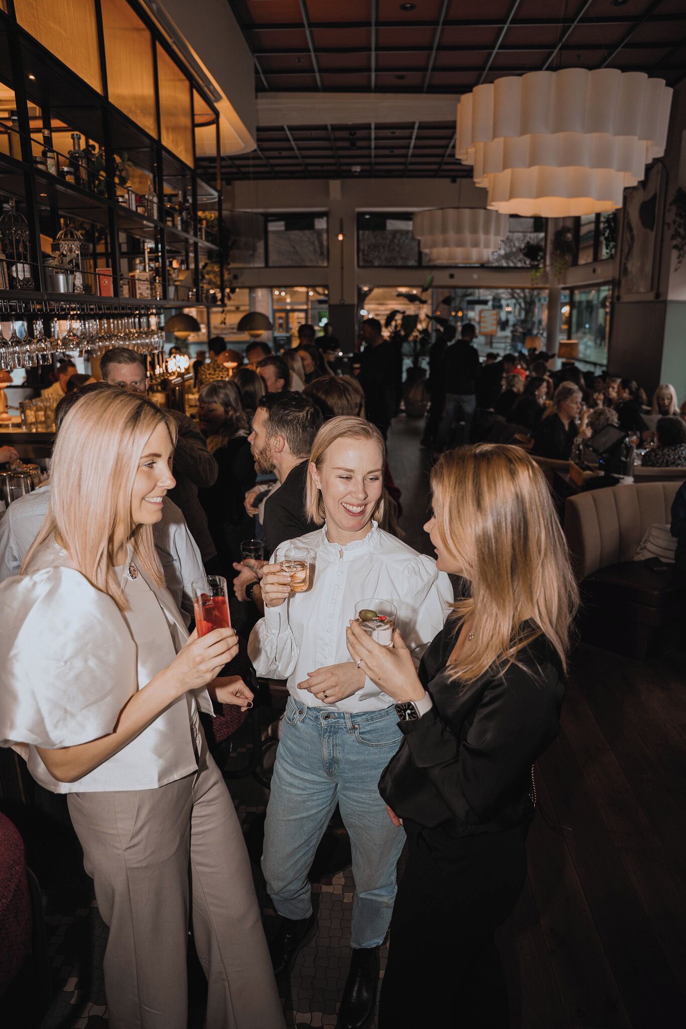 Zona de bar con estantes llenos de botellas y vasos, una lámpara colgante y gente conversando.