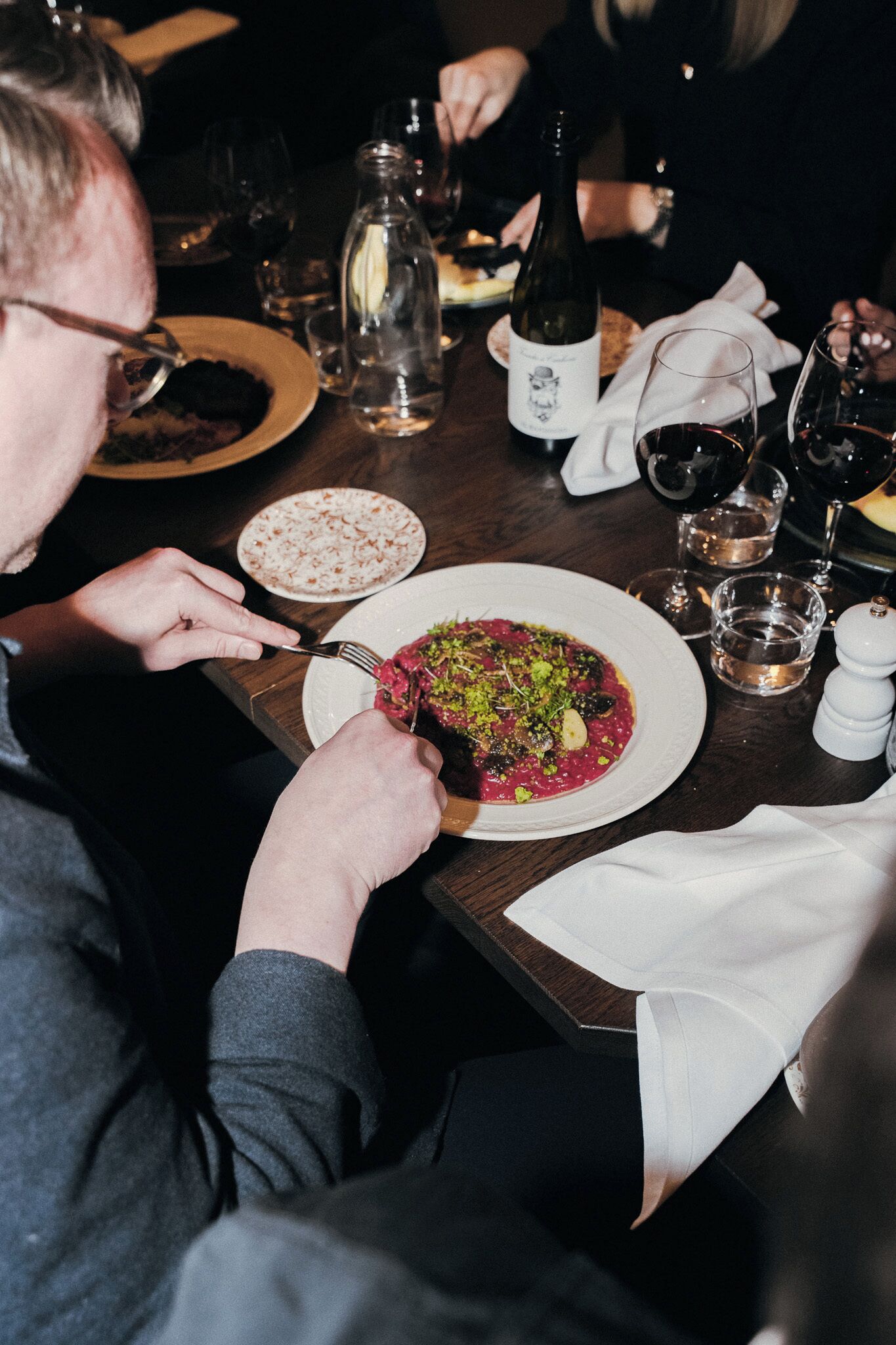 Una persona cenando en una mesa con un plato de comida, una botella de vino y copas.
