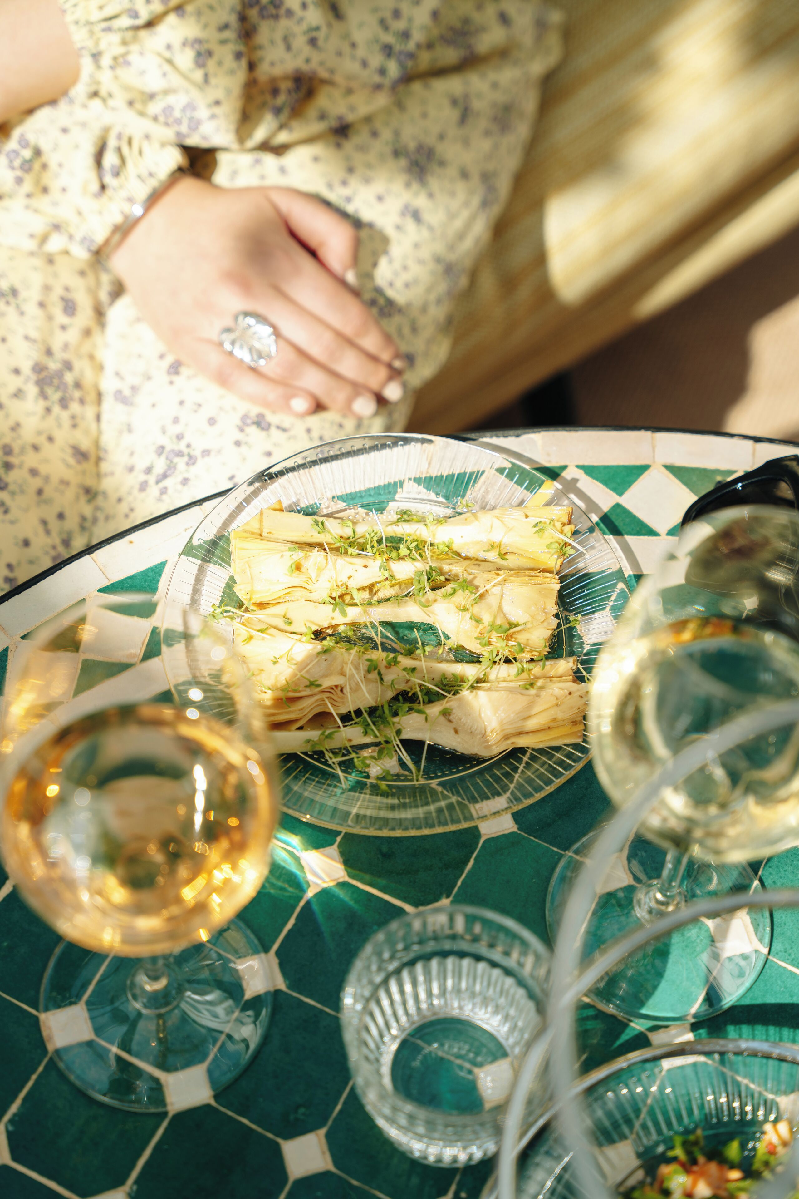 Una mesa con un plato de comida bajo un vidrio, copas de vino y un mantel decorativo.