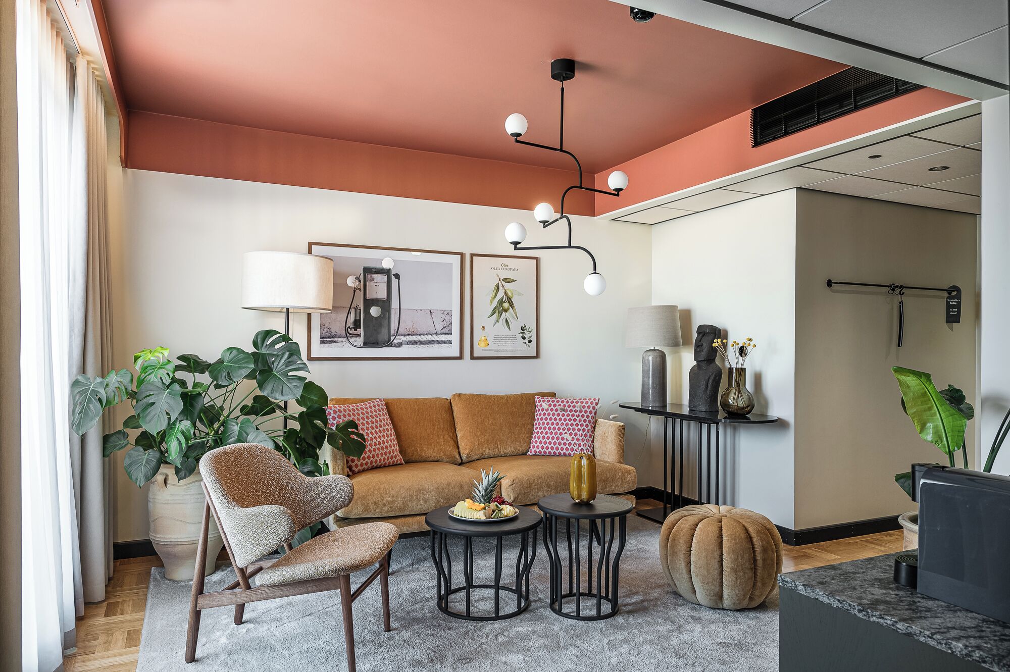 A modern living room with a brown sofa, a patterned armchair, and a round ottoman. There are two small round tables, one with a fruit bowl and the other with a vase. A large potted plant is on the left, and a wall-mounted shelf with decorative items is on the right.