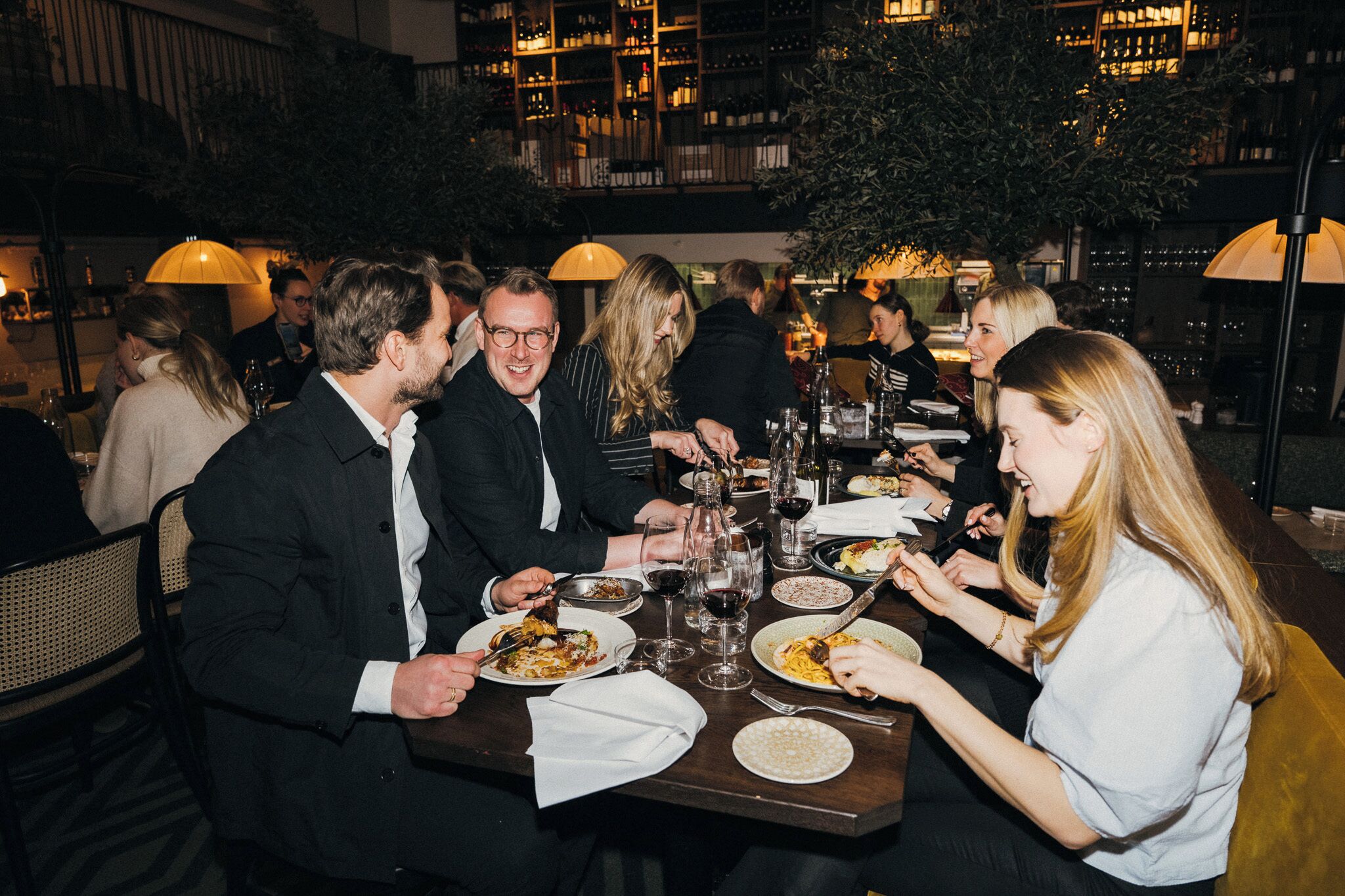 A group of people dining at an indoor restaurant with tables set for meals and wine glasses.