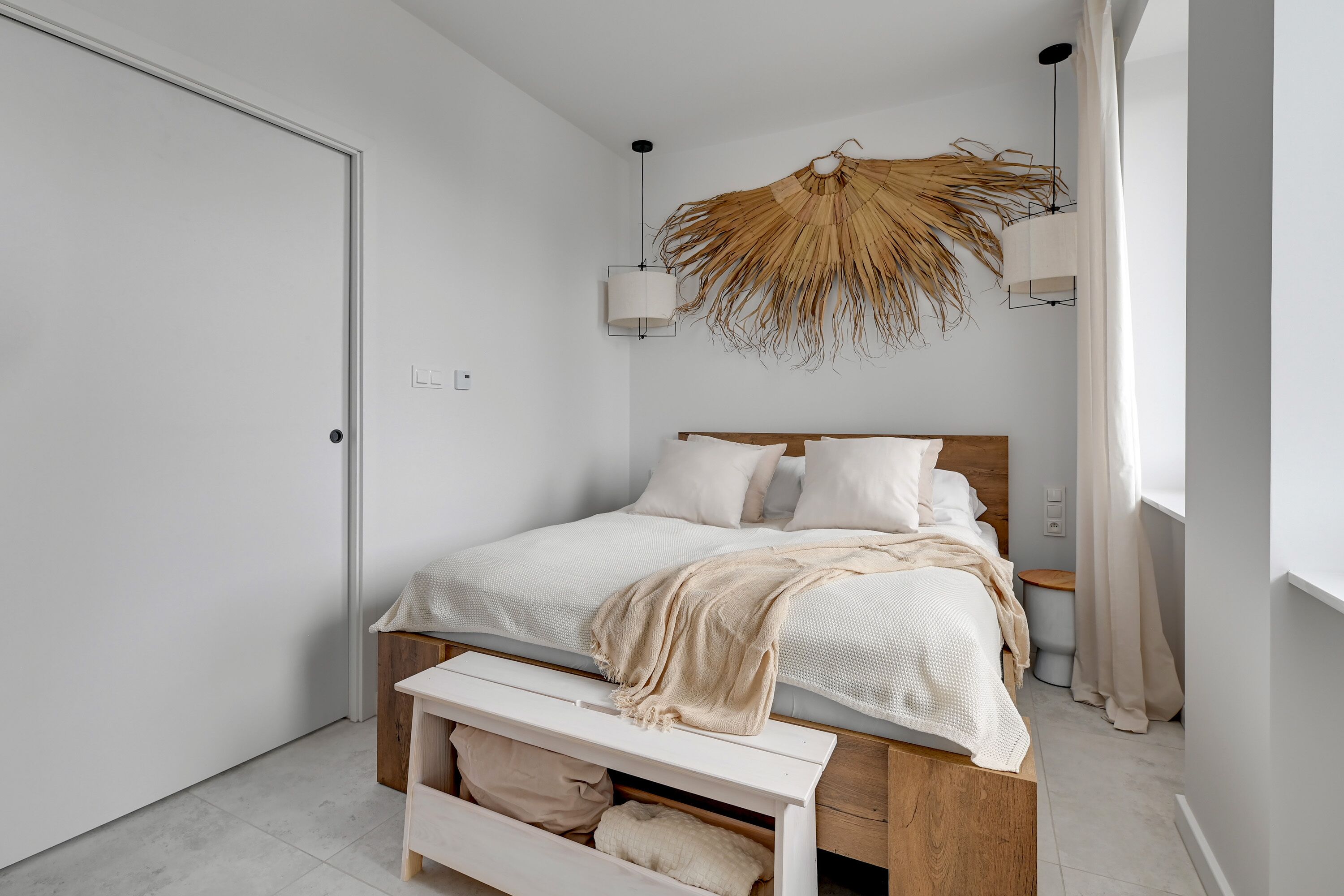A modern bedroom with a bed, bedside tables, a bench, a wall-mounted light fixture, and a door.