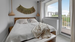 A bedroom with a large window, a bed with white bedding, a woven wall hanging, and two hanging lamps.