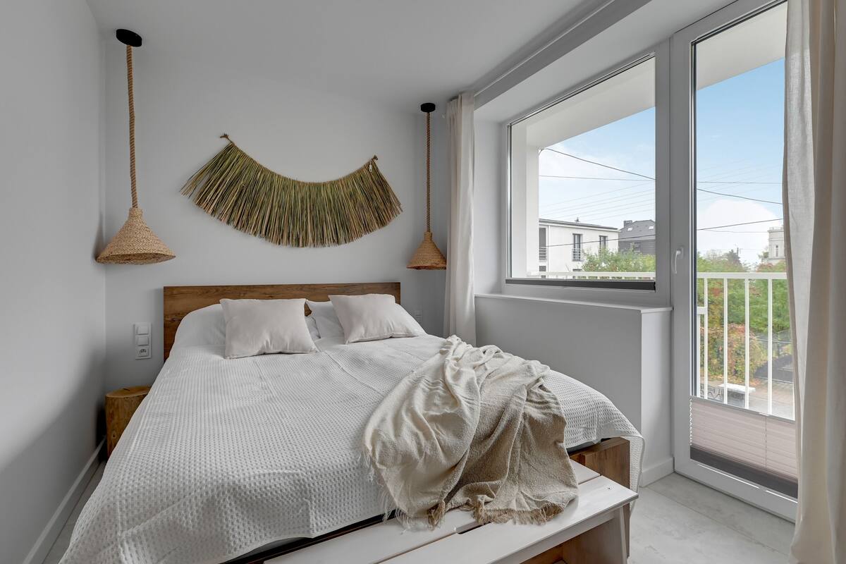 Een slaapkamer met een groot raam, een bed met wit beddengoed, een wandkleed en twee hanglampen.
