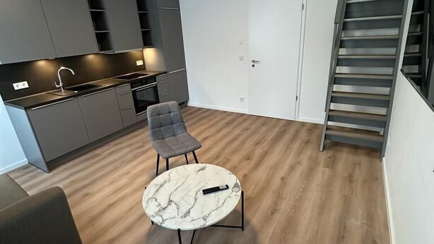 Et moderne køkken med lysegrå skabe, indbygget ovn og vask. Der er en spiseplads med et marmorbord og to grå stole. En trappe er synlig i baggrunden.
