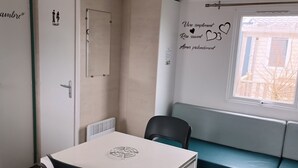 Ett litet, modernt rum med ett vitt bord och två stolar, en bänk med turkosa kuddar, ett fönster med utsikt över en byggnad och en vägg med en skylt som visar var toaletten finns samt ett dekorativt citat.