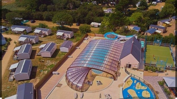 Ett flygbildsfoto av ett semesterområde med flera stugor, en stor pool under tak och en lekplats för barn.