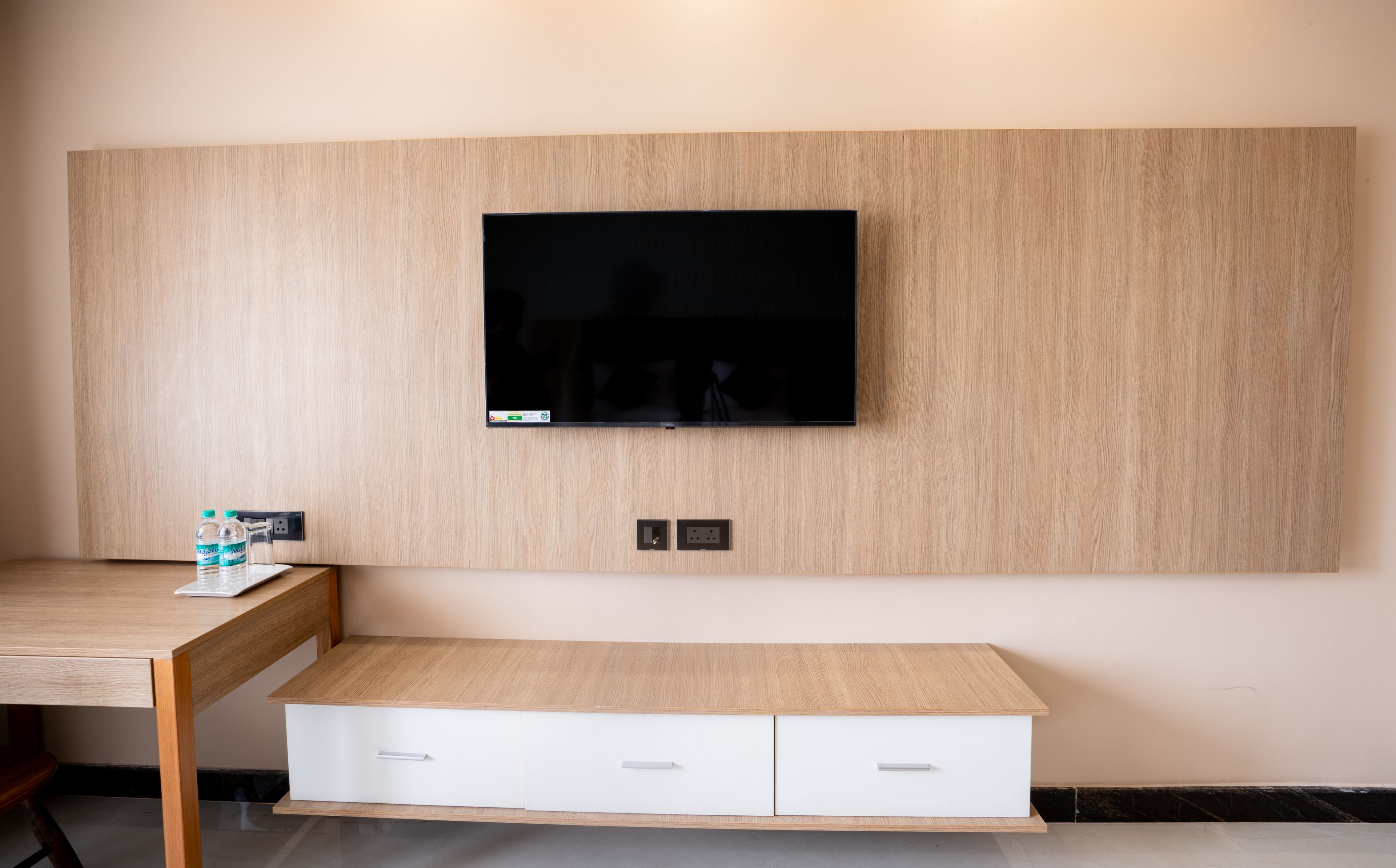 A modern room with a flat-screen TV mounted on a wooden wall, a wooden desk with a chair, and a white bench with drawers.