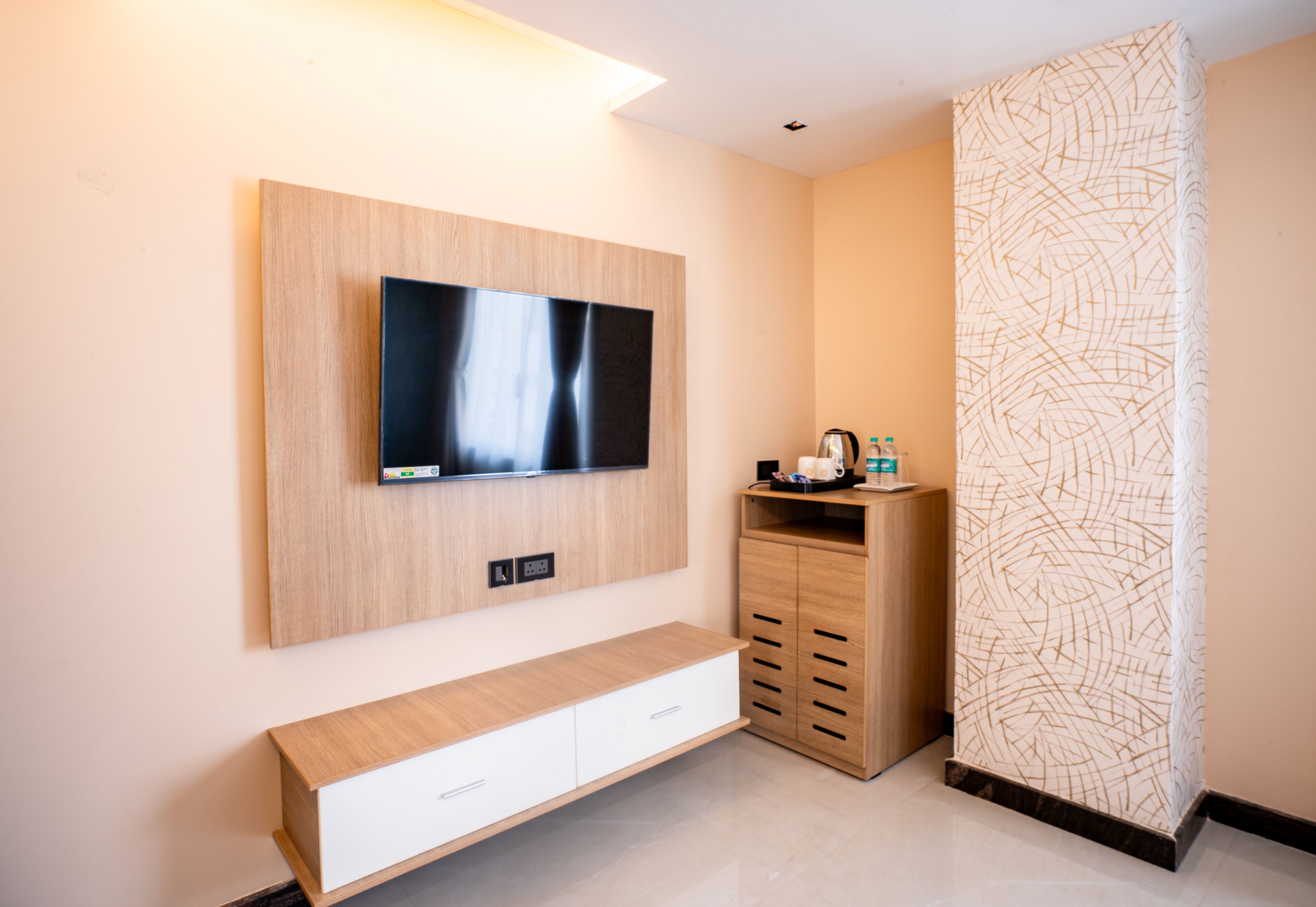 Ein modernes Hotelzimmer mit einem flachbildfernseher, der an einer Holzvertäfelung angebracht ist, einem weißen Konsolentisch mit Schubladen, einem Holzschrank mit einem gemusterten Vorhang und einem gefliesten Boden.