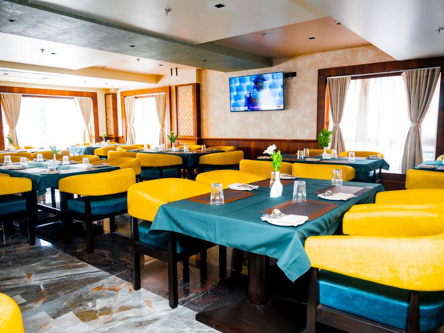 Un comedor con sillas tapizadas en amarillo y azul, manteles verdes y un televisor de pantalla plana montado en la pared.
