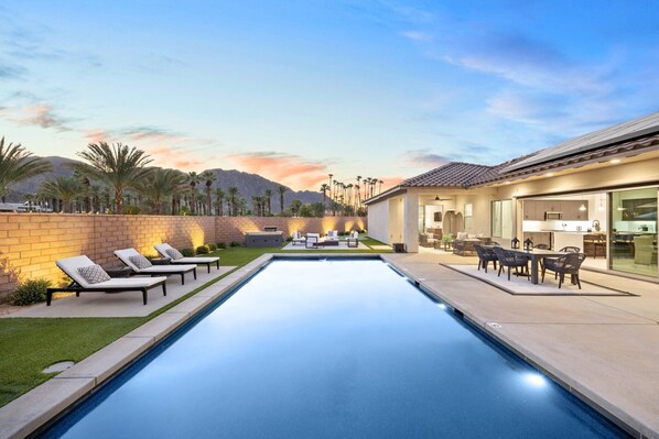 A modern outdoor pool area with lounge chairs, a barbecue, and a patio with a kitchen visible in the background. - Vista Santa Rosa: Pool, Spa, Firepit, BBQ, EV Charger (La Quinta)