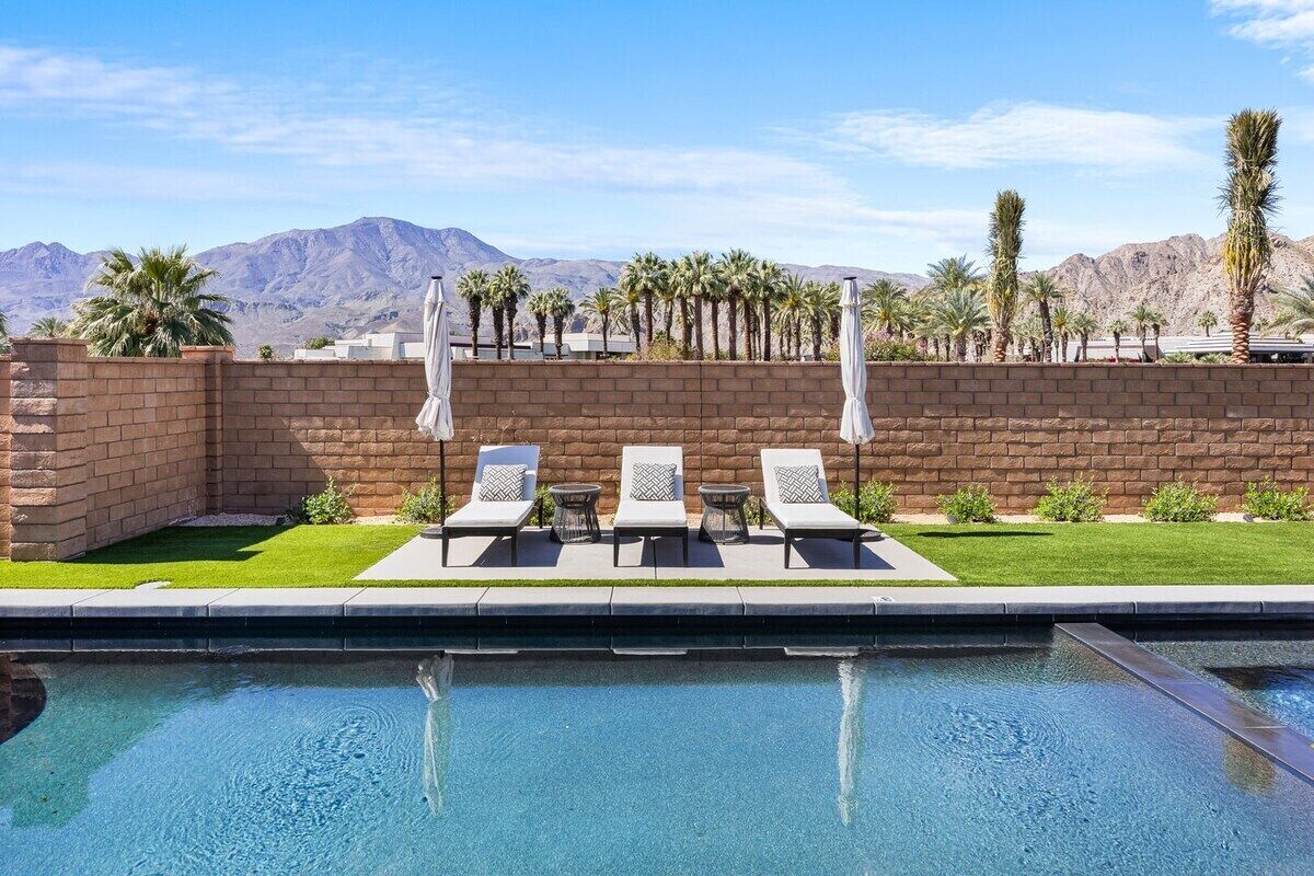 A swimming pool with lounge chairs and umbrellas, surrounded by a brick wall and palm trees, with mountains in the background.