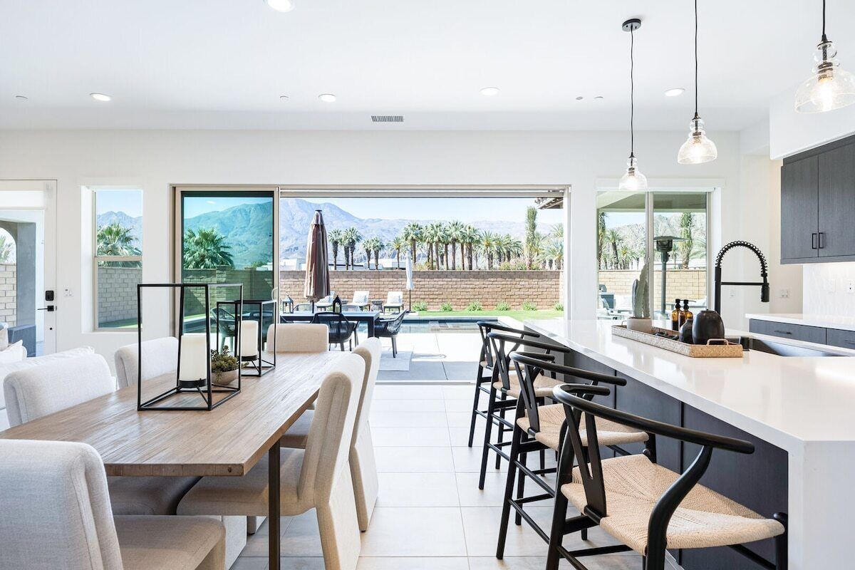 Une cuisine moderne dotée d’un grand îlot central, d’un coin repas avec une table et des chaises en bois, et offrant une vue sur un paysage montagneux grâce à de grandes portes coulissantes.