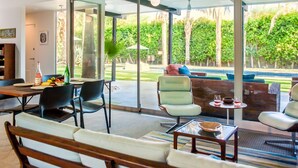 A modern living room with a large glass door leading to a patio area. There is a dining table with chairs, a sofa, and a coffee table.