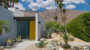 A modern building with a yellow door, surrounded by a landscaped garden with rocks and plants, and mountains in the background.