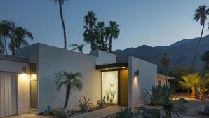 A modern house with a well-lit entrance, surrounded by palm trees and rocks.