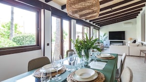 Uma sala de jantar com uma mesa posta para quatro pessoas, grandes janelas, uma luminária de teto trançada e uma área de estar moderna com televisão de tela plana.