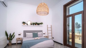 Quarto com cama, luminária pendente, janela e vista para a praia.