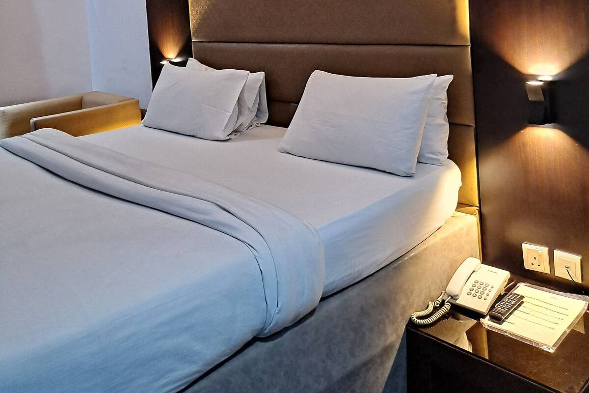 A neatly made bed with white linens, a wooden nightstand with a telephone and a notepad, and a wall-mounted light fixture.