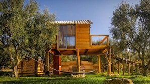Une cabane en bois perchée dans un arbre, dotée d’une porte en verre et entourée d’une clôture en corde, située dans une zone herbeuse bordée d’arbres.