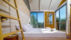 Une chambre avec un mur en lambris de bois, un lit recouvert de draps blancs, une échelle en bois et de grandes fenêtres donnant sur la forêt.