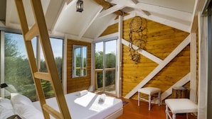 Une chambre à coucher avec un plafond en bois, un lit, deux chaises et de grandes fenêtres donnant sur la forêt.