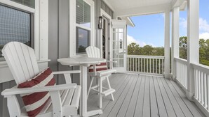 A covered porch with white chairs and a table, a window with a view of trees, and a white railing. - Charming 3BR Home on 30A – Golf Cart Included, 5 Min to Beach (Santa Rosa Beach)