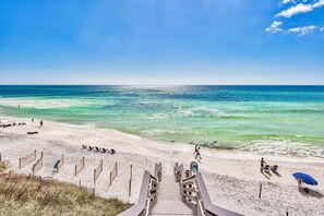 Une plage avec un escalier en bois menant au rivage, une eau turquoise et limpide, et quelques personnes sur le sable.