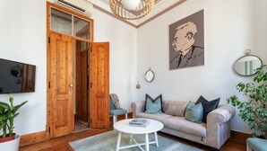 En stue med en beige sofa, et hvidt sofabord, en trædør, et fjernsyn og en potteplante.
