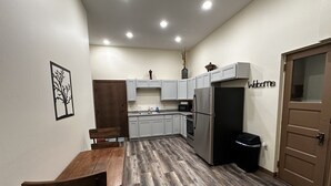 A compact kitchen with white cabinets, a stainless steel refrigerator, a wooden dining table, and a dark trash bin.