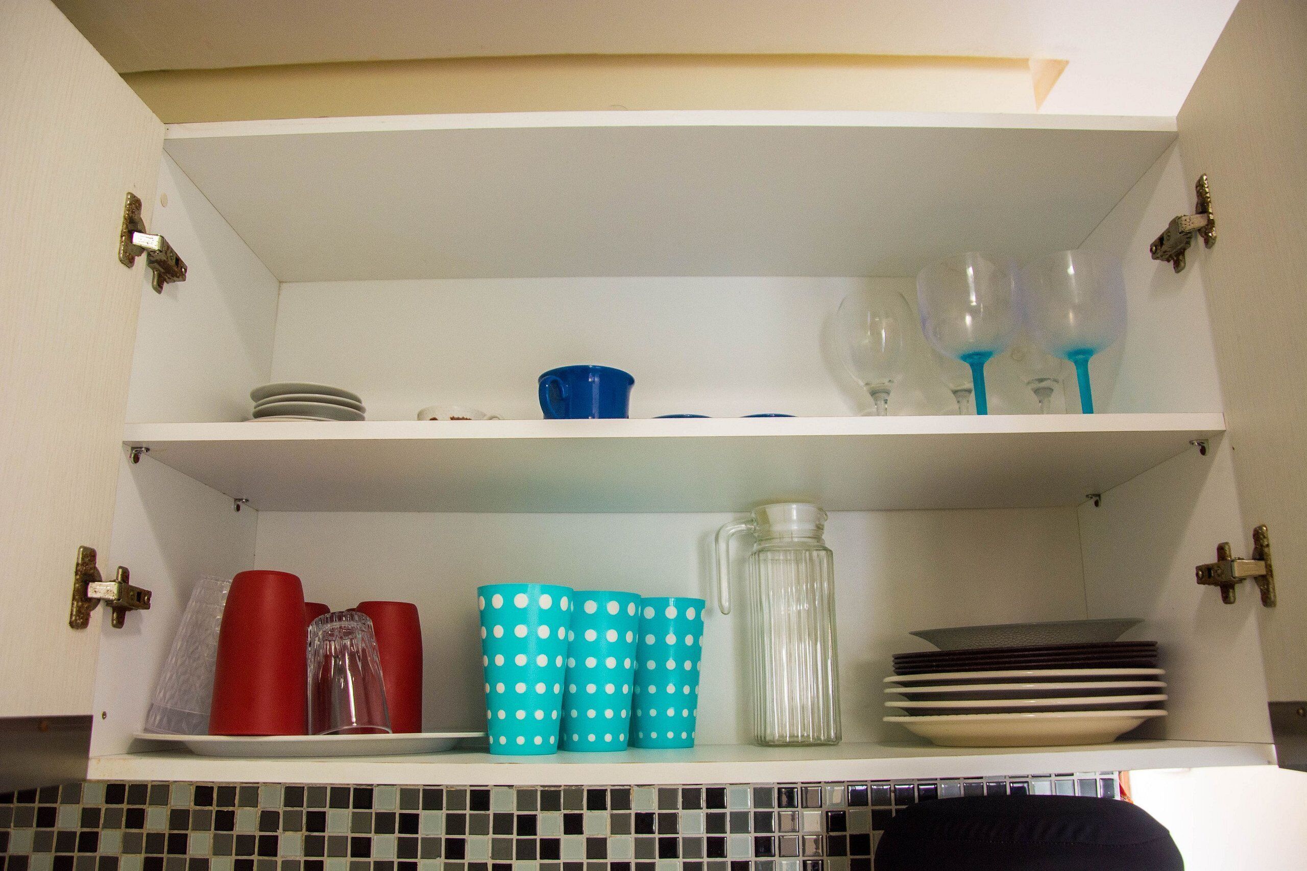 Uma cozinha com armários brancos, painel de azulejos e prateleiras com diversos utensílios de cozinha, incluindo xícaras, pratos e copos.