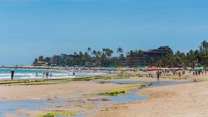 A beach with a large building in the background, palm trees, and people walking. - Manawa Beach Flat by All (Ipojuca)