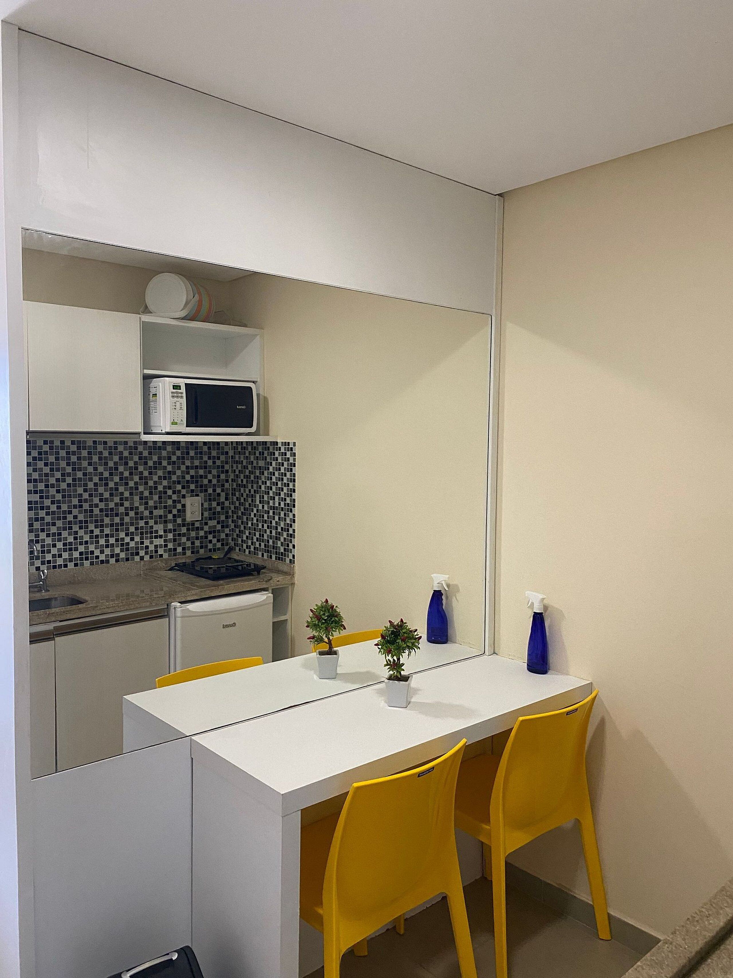 Cocina compacta con microondas, pileta y salpicadero de azulejos. Posee una pequeña mesa de comedor con dos sillas amarillas y dos plantas en macetas.