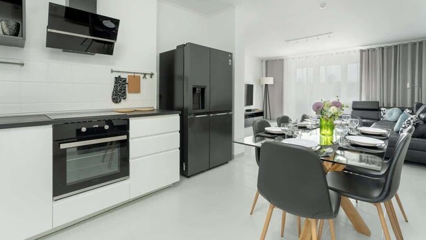 Eine moderne Küche mit weißen Schränken, einem schwarzen Kühlschrank und einem Essbereich mit einem Glastisch und schwarzen Stühlen.