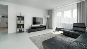 Ein modernes Wohnzimmer mit einer schwarzen Ledersofa, einem Flachbildfernseher, einem Bücherregal und einem großen Fenster mit transparenten Vorhängen.
